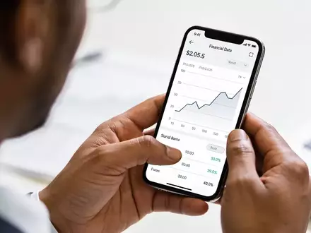 Nahaufnahme einer Hand, die ein Smartphone mit Finanzdiagrammen in einer professionellen Büroumgebung hält. - Foto: über boerse-global.de