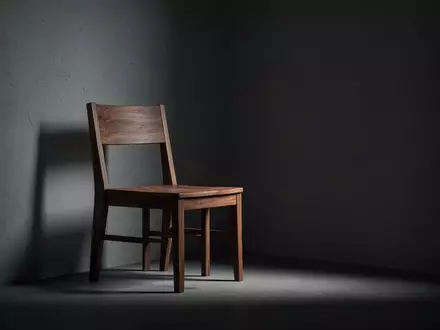 Ein leerer Holzstuhl in einem schattigen, minimalistischen Raum symbolisiert die Isolation und Verletzlichkeit durch Betrug. - Foto: über boerse-global.de
