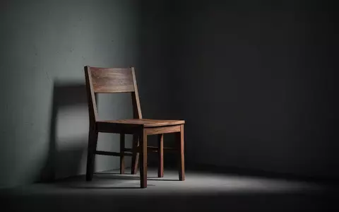 Ein leerer Holzstuhl in einem schattigen, minimalistischen Raum symbolisiert die Isolation und Verletzlichkeit durch Betrug. - Foto: über boerse-global.de