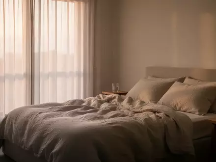 Ein minimalistisch eingerichtetes Schlafzimmer in sanftem Morgenlicht mit einem ungemachten Bett und einem Glas Wasser. - Foto: über boerse-global.de