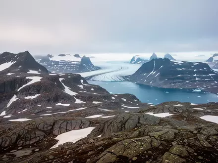 Weite arktische Landschaft in Grönland mit felsigen Bergen, Schnee und einem tiefblauen Fjord unter bewölktem Himmel. - Foto: ĂŒber boerse-global.de