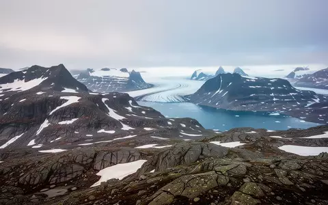 Weite arktische Landschaft in Grönland mit felsigen Bergen, Schnee und einem tiefblauen Fjord unter bewölktem Himmel. - Foto: über boerse-global.de