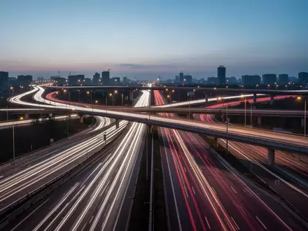 Eine mehrspurige Autobahnkreuzung bei Dämmerung mit Lichtspuren vorbeifahrender Fahrzeuge in einer urbanen Landschaft. - Foto: über boerse-global.de