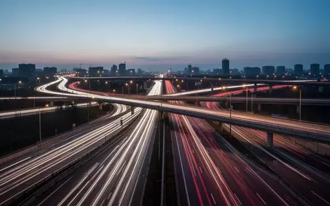Eine mehrspurige Autobahnkreuzung bei Dämmerung mit Lichtspuren vorbeifahrender Fahrzeuge in einer urbanen Landschaft. - Foto: über boerse-global.de