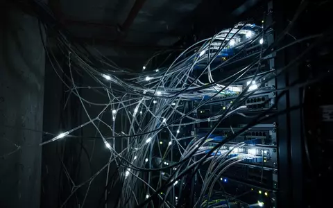 Ein dunkler, technischer Hintergrund mit verschlungenen Glasfaserkabeln und schwachen Lichtquellen, der eine digitale Bedrohung symbolisiert. - Foto: über boerse-global.de