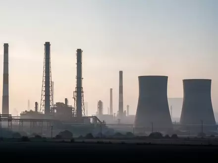Weitwinkelaufnahme einer industriellen Anlage mit Schornsteinen und Kühltürmen in einer weitläufigen Landschaft bei Dämmerung. - Foto: über boerse-global.de