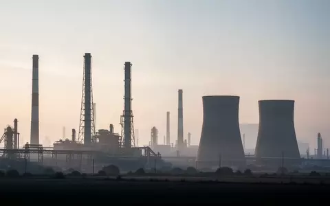 Weitwinkelaufnahme einer industriellen Anlage mit Schornsteinen und Kühltürmen in einer weitläufigen Landschaft bei Dämmerung. - Foto: über boerse-global.de
