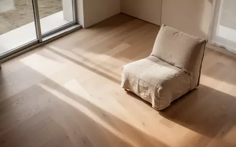 Ein minimalistisch eingerichteter, leerer Raum mit hellem Holzboden und einem einzelnen beigen Leinenstuhl, der durch einfallendes Sonnenlicht und Schatten geprägt ist. - Foto: über boerse-global.de