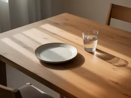 Ein schlichter, sonnendurchfluteter Esstisch mit einem leeren Teller und einem Glas Wasser in einer ruhigen, minimalistischen Umgebung. - Foto: über boerse-global.de