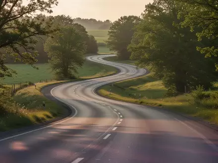 Eine menschenleere, kurvige Landstraße in einer grünen Hügellandschaft bei spätem Nachmittagslicht. - Foto: über boerse-global.de
