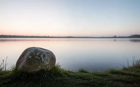 Ein ruhiger See bei Sonnenaufgang mit einem einzelnen Stein am Ufer als Symbol für Stille und Reflexion. - Foto: über boerse-global.de