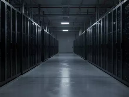 Dunkler, weitläufiger Serverraum mit metallenen Regalsystemen und reflektierendem Boden in einer kühlen, industriellen Atmosphäre. - Foto: über boerse-global.de