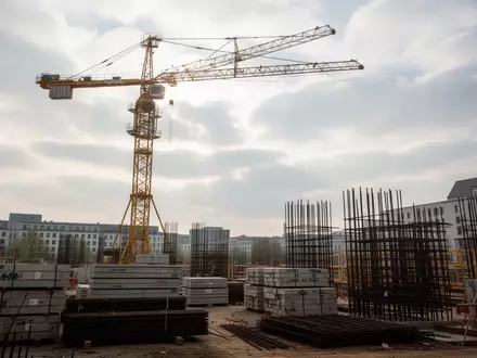 Eine Baustelle mit einem gelben Baukran vor einem bewölkten Himmel in Berlin, umgeben von Baumaterialien. - Foto: über boerse-global.de
