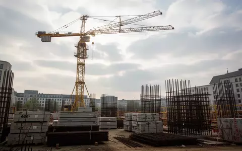 Eine Baustelle mit einem gelben Baukran vor einem bewölkten Himmel in Berlin, umgeben von Baumaterialien. - Foto: über boerse-global.de