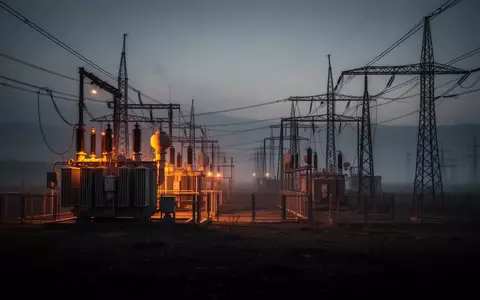 Ein dunkles, industrielles Stromnetz-Gelände bei Dämmerung mit glühenden Transformatoren und Hochspannungsleitungen in einer nebligen Landschaft. - Foto: über boerse-global.de