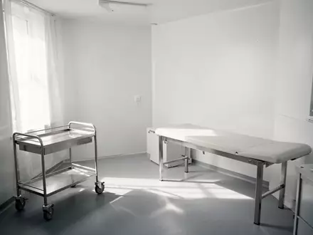 Ein minimalistischer, leerer Behandlungsraum in einer Arztpraxis mit einem Edelstahltrolley und einer Untersuchungsliege bei natürlichem Tageslicht. - Foto: über boerse-global.de