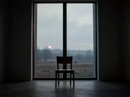 Ein einsamer Stuhl vor einem Fenster in einem schattigen Raum bei Dämmerung, das eine ruhige Landschaft überblickt. - Foto: über boerse-global.de