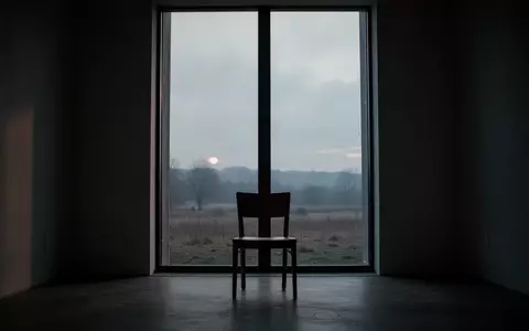 Ein einsamer Stuhl vor einem Fenster in einem schattigen Raum bei Dämmerung, das eine ruhige Landschaft überblickt. - Foto: über boerse-global.de