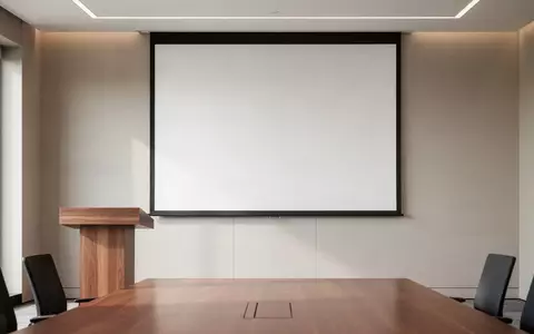 Ein minimalistischer Konferenzraum mit einem leeren Rednerpult vor einer unbeschriebenen, weißen Projektionsfläche. - Foto: über boerse-global.de