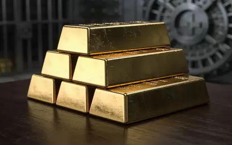 Mehrere glänzende Goldbarren, die auf einem dunklen Holztisch in einem Tresorraum gestapelt sind. - Foto: über boerse-global.de
