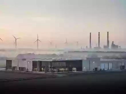 Ein modernes Industriegebäude in einer ruhigen Landschaft mit Windrädern im Hintergrund bei Dämmerung. - Foto: über boerse-global.de