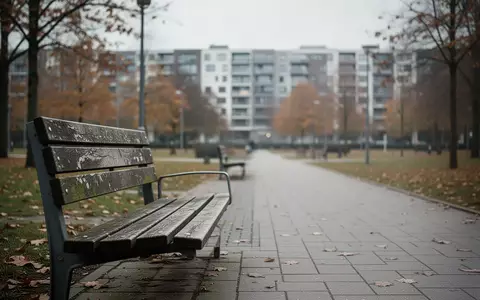 Ein leerer Parkweg mit einer Parkbank und im Hintergrund unscharf erkennbare Wohngebäude bei bewölktem Tageslicht. - Foto: über boerse-global.de