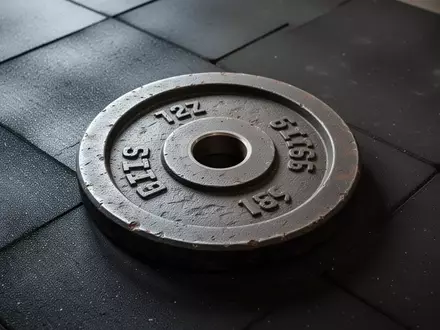 Eine schwere, texturierte Hantelscheibe liegt auf einem dunklen Gummiboden in einem Fitnessraum. - Foto: über boerse-global.de