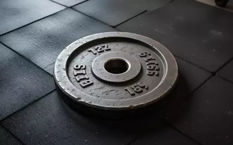 Eine schwere, texturierte Hantelscheibe liegt auf einem dunklen Gummiboden in einem Fitnessraum. - Foto: über boerse-global.de