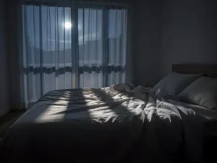 Ein minimalistisch eingerichtetes Schlafzimmer in der Nacht mit sanftem Mondlicht, das durch Vorhänge auf ein ungemachtes Bett fällt. - Foto: über boerse-global.de