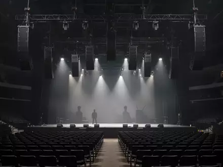 Ein professionelles Line-Array-Lautsprechersystem, das in einer dunklen Konzerthalle von der Decke hängt. - Foto: über boerse-global.de