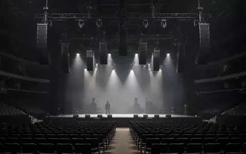 Ein professionelles Line-Array-Lautsprechersystem, das in einer dunklen Konzerthalle von der Decke hängt. - Foto: über boerse-global.de