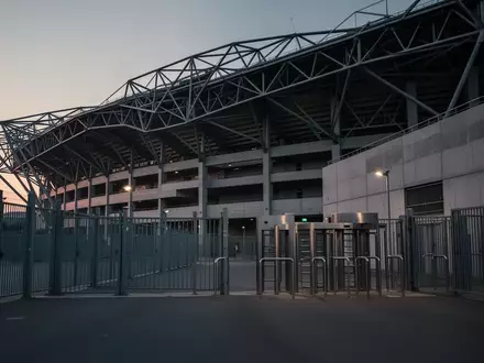 Die Außenfassade eines leeren Fußballstadions in der Abenddämmerung mit Fokus auf Architektur und Metallzäune. - Foto: über boerse-global.de