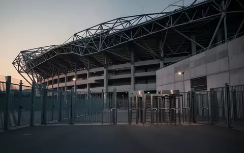 Die Außenfassade eines leeren Fußballstadions in der Abenddämmerung mit Fokus auf Architektur und Metallzäune. - Foto: über boerse-global.de