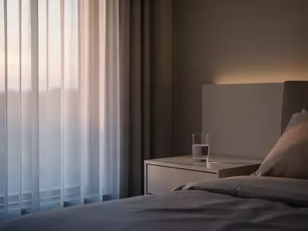 Ein minimalistisch eingerichtetes Schlafzimmer in den frühen Morgenstunden mit sanftem Tageslicht, das durch einen Vorhang fällt. - Foto: über boerse-global.de
