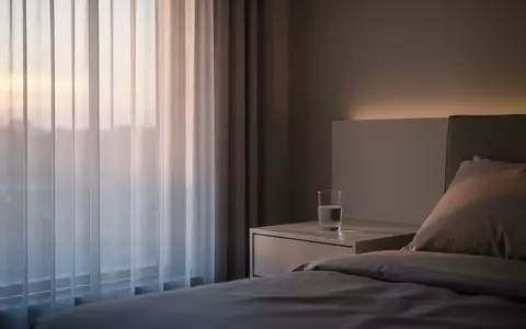 Ein minimalistisch eingerichtetes Schlafzimmer in den frühen Morgenstunden mit sanftem Tageslicht, das durch einen Vorhang fällt. - Foto: über boerse-global.de