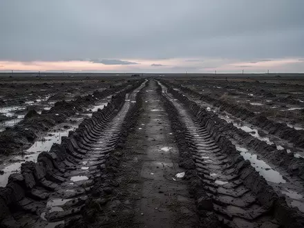 Ein rauer, schlammiger Feldweg mit tiefen Reifenspuren in einer weiten, unbewohnten Landschaft bei Dämmerlicht. - Foto: über boerse-global.de