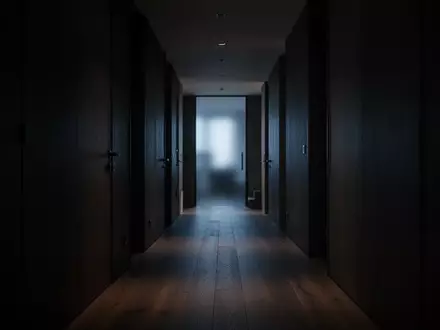 Ein dunkler, leerer Flur in einer modernen Wohnung bei Nacht, der auf eine entfernte, schwach beleuchtete Tür zuführt. - Foto: über boerse-global.de