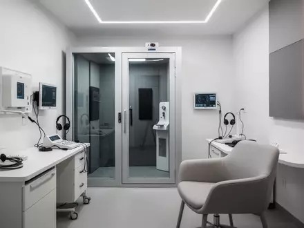 Ein minimalistisch eingerichteter, moderner Audiometrie-Raum mit einer schallisolierten Prüfkabine in einer klinischen Umgebung. - Foto: über boerse-global.de