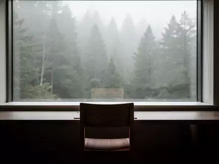 Ein minimalistischer, ruhiger Arbeitsraum mit einem leeren Stuhl vor einem Fenster, das auf einen nebligen Wald blickt. - Foto: über boerse-global.de