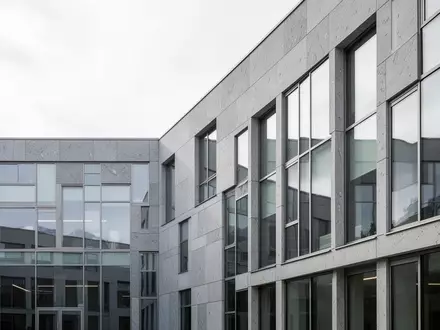 Moderne Architektur eines Verwaltungsgebäudes mit grauer Steinfassade und Glasfront unter bewölktem Himmel. - Foto: über boerse-global.de