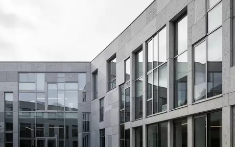 Moderne Architektur eines Verwaltungsgebäudes mit grauer Steinfassade und Glasfront unter bewölktem Himmel. - Foto: über boerse-global.de