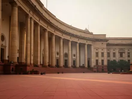 Die Fassade eines neoklassizistischen Regierungsgebäudes in New Delhi bei goldenem Licht mit großen Steinsäulen und einem leeren Vorplatz. - Foto: über boerse-global.de