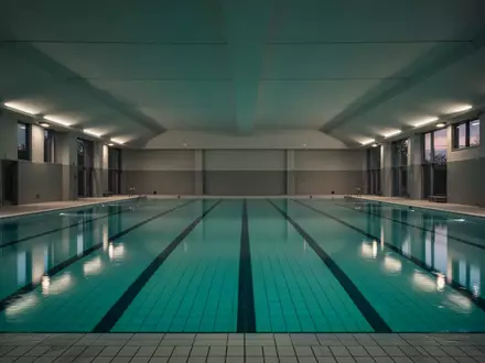 Ein leeres Hallenbad mit ruhigem, blauem Wasser und architektonischen Linien bei gedimmter Beleuchtung. - Foto: über boerse-global.de