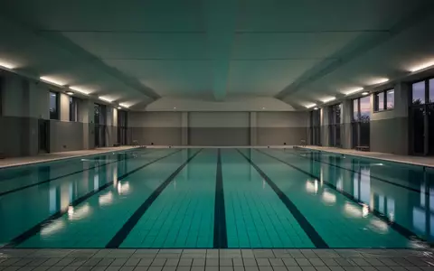 Ein leeres Hallenbad mit ruhigem, blauem Wasser und architektonischen Linien bei gedimmter Beleuchtung. - Foto: über boerse-global.de