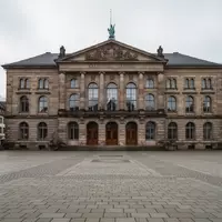 Ein historisches Rathausgebäude mit einem leeren Platz davor bei bewölktem Tageslicht. - Foto: über boerse-global.de