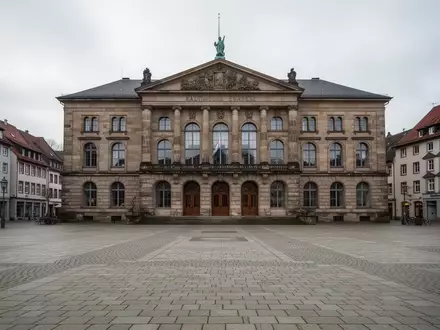 Ein historisches Rathausgebäude mit einem leeren Platz davor bei bewölktem Tageslicht. - Foto: über boerse-global.de