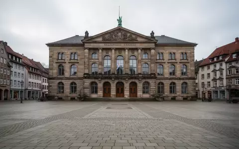 Ein historisches Rathausgebäude mit einem leeren Platz davor bei bewölktem Tageslicht. - Foto: über boerse-global.de