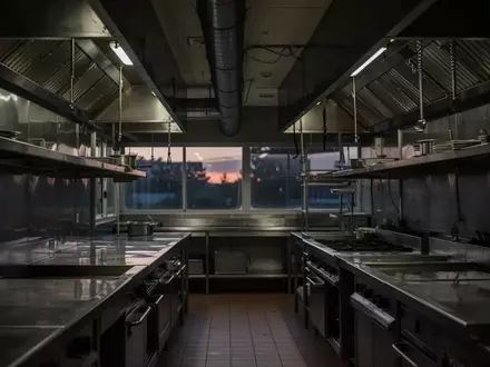 Leere, professionelle Gastronomieküche mit Edelstahl-Oberflächen und Abzugshauben in einer schummrigen Lichtstimmung. - Foto: über boerse-global.de