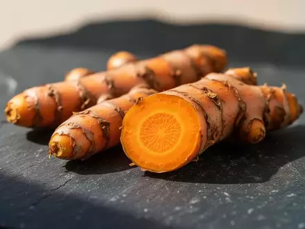 Frische Kurkumawurzeln auf einer dunklen Steinoberfläche, wobei eine Wurzel aufgeschnitten ist und ihr leuchtend gelbes Inneres zeigt. - Foto: über boerse-global.de