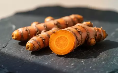 Frische Kurkumawurzeln auf einer dunklen Steinoberfläche, wobei eine Wurzel aufgeschnitten ist und ihr leuchtend gelbes Inneres zeigt. - Foto: über boerse-global.de